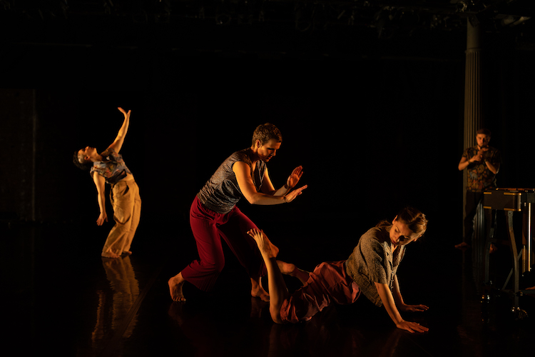 in dim light one dancer in loose fitting pants and a midriff top stands in profile while lifting one arm to the ceiling, in front of her are two other dancers in grey tops of varied sleeve length and dark pants,one dancer attempts to pat the back of another dancer whose lower body lays on the floor, while her upper body arches and twists supported by her arms, in the very back is a musician playing a flute